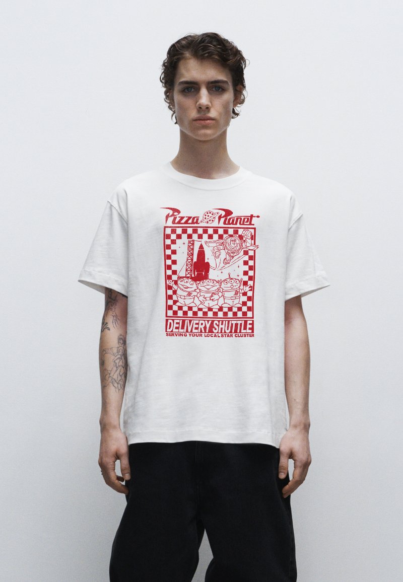 Bílá bavlněná tričko s červeným potiskem a kostkovaným okrajem, textem "Pizza Planet" a kreslenými postavičkami. Volný střih, krátké rukávy.