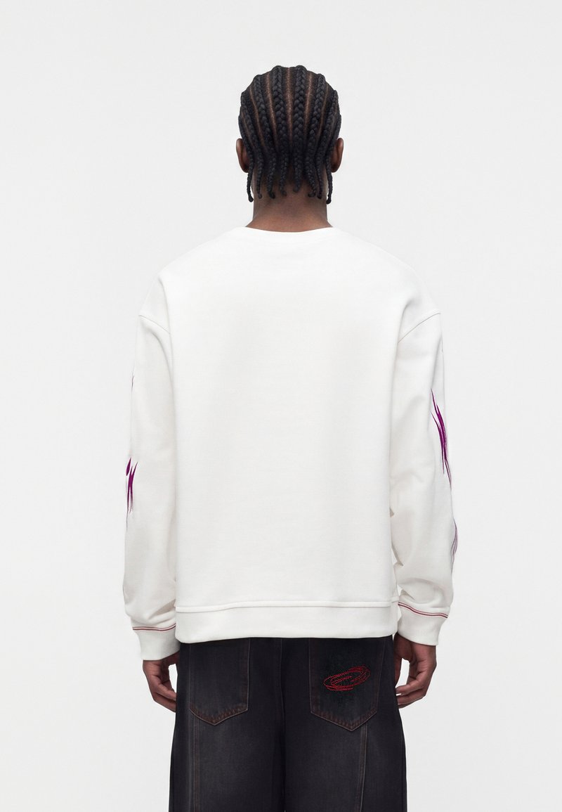 Person with braided hair facing away wearing a white long-sleeve top with purple details on sleeves and black pants with red stitching.