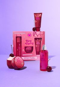 Un imballaggio rosa con illustrazioni di ciliegie circonda cinque prodotti di bellezza: lozione, profumo per il corpo, scrub e crema, su uno sfondo viola.
