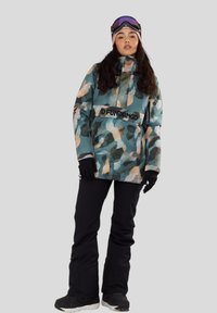 Žena v tyrkysové a broskvové kamuflážové sněhové bundě, černých sněhových kalhotách, rukavicích, lyžařských brýlích a čepici, stojící na jednobarevném pozadí.