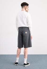 Pantalones cortos de denim gris con un ajuste relajado y un bolsillo trasero que presenta un logo de Carhartt, combinados con una camiseta de manga larga blanca y zapatillas.