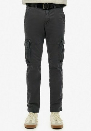 Pantalon cargo gris foncé avec poches latérales, ceinture noire, porté avec des baskets beige et blanc, vu de la taille jusqu'aux pieds sur fond blanc.