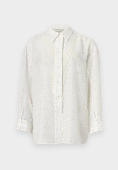 Chemise blanche à manches longues avec un tissu léger, présentant un motif texturé subtil et de petits sequins réfléchissants répartis sur toute la surface. Manches longues avec poignets.