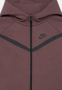 Hrdzavohnedá mikina na zips s čiernym zipsom, logom Nike a čiernym akcentom na prsiach. Hladká, štruktúrovaná látka s kapucňou.
