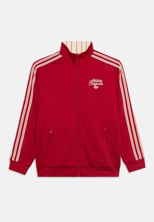 Veste de survêtement rouge Adidas avec trois bandes blanches sur les manches, fermeture éclair à l'avant, poches zippées et logo « Adidas Originals » sur la poitrine.