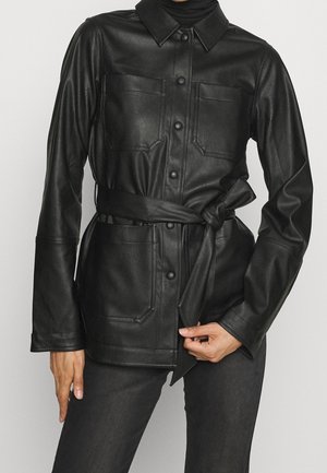 Chaqueta de cuero sintético negro con cinturón, botones a presión en el frente, bolsillos en el pecho y cadera, llevada sobre pantalones negros.