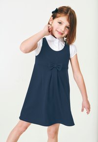 Navyblaues A-Linien-Kleid, ärmellos mit einer Schleifenakzent am Bund, kombiniert mit einem weißen Hemd mit Kragen darunter. Glatte Stofftextur.