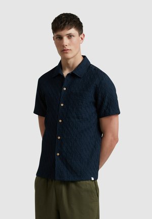 Marineblauw shirt met korte mouwen en een gestructureerd patroon, voorzien van een kraag, een borstzak en houten knopen. Gecombineerd met groene broek.