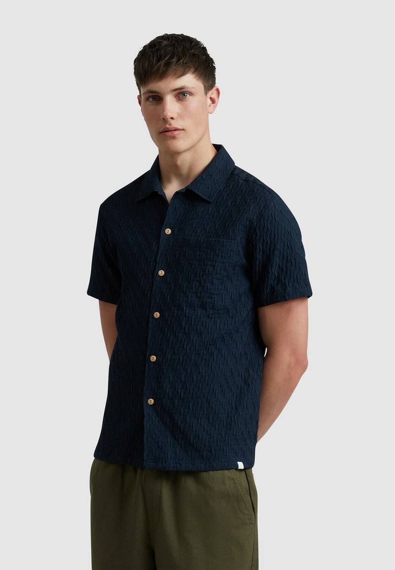 Chemise courte à manches en bleu marine avec un motif texturé, dotée d'un col, d'une poche poitrine et de boutons en bois. Associée à un pantalon vert.