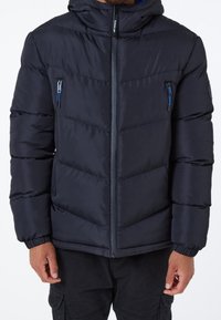 Uomo che indossa un piumino nero con cappuccio, zip frontale e tasche sul petto con zip, abbinato a pantaloni cargo neri.