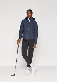 Marinblå vindjacka med dragkedja, huva och framfickor; kombinerad med svarta byxor och vita sneakers, hållande en golfklubba.