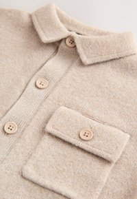 Chemise en tricot beige avec un col chemise, une fermeture à boutons et une poche poitrine. Présente des boutons ronds de couleur noisette et une texture douce.