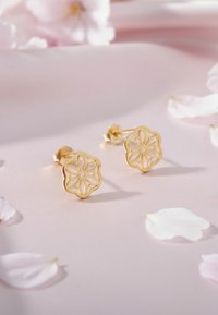 Guldblommiga örhängen med intrikata utskärningar. Lagt på en mjuk rosa yta med spridda vita blombladsprydnader.