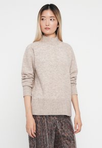 Beige neulepaita, jossa on korkea kaula-aukko, ribbireunukset ja -helma. Pehmeä rakenne, hieman ylisuuri istuvuus ja vähäiset sauman yksityiskohdat.