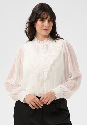 Femme aux cheveux foncés portant un chemisier blanc transparent à volants et un pantalon noir, debout les mains jointes devant elle.