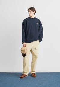 Μπλε σκούρα φούτερ με λογότυπο, μπεζ παντελόνια cargo και καφέ παπούτσια· κρατώντας μια ανοιχτό καφέ καπέλο· απαλό ύφασμα, χαλαρή εφαρμογή, μινιμαλιστικός σχεδιασμός.