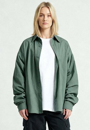 Giovane con capelli biondi fino alle spalle indossa una camicia verde oversize con bottoni sopra una t-shirt bianca e pantaloni cargo neri, in piedi davanti a uno sfondo semplice.