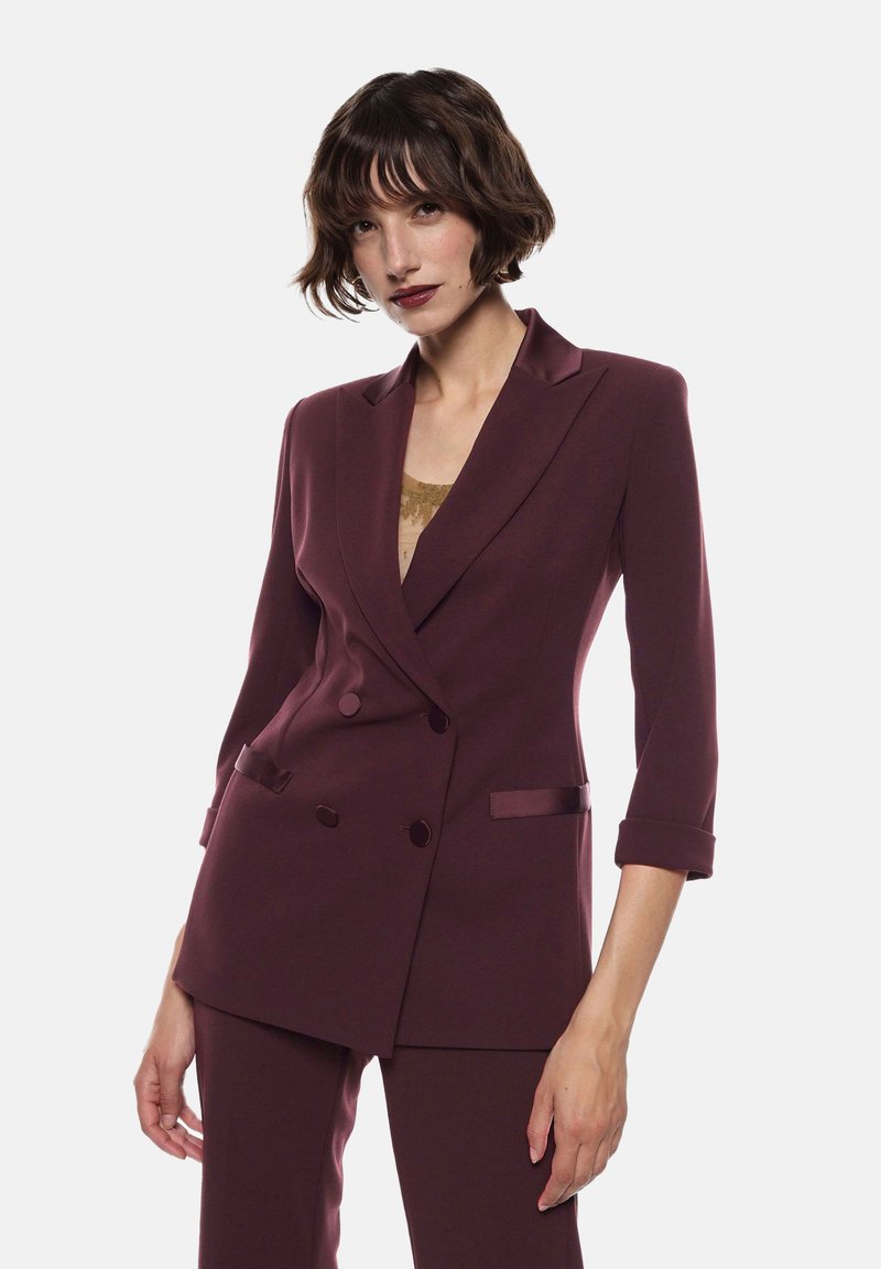 Blazer bordeaux doppiopetto con rifiniture in satin, rever a punta, maniche 3/4 e vestibilità sartoriale. Indossato sopra un top dorato.