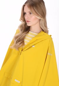 Pelerină de ploaie galbenă impermeabilă cu glugă, închideri prin capse și detaliu cu logo pe mânecă, împreună cu o cămașă galbenă cu dungi dedesubt.
