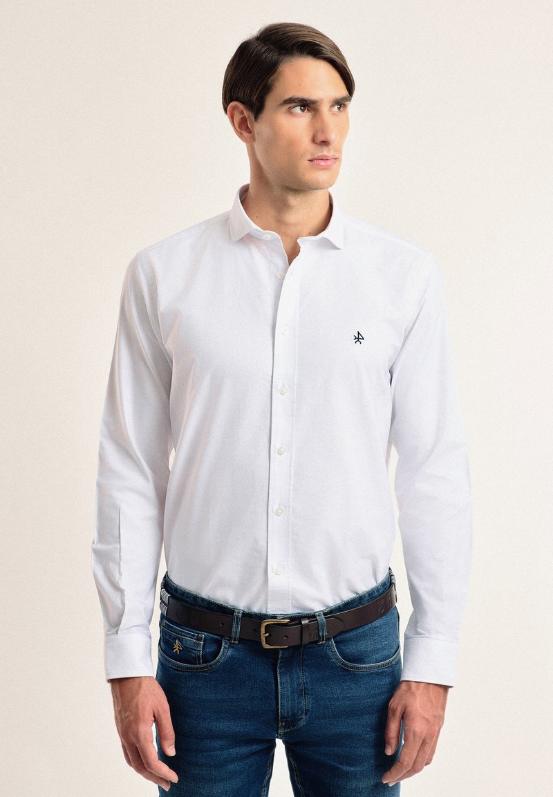 Camisa blanca de botones hecha de tela suave, con mangas largas y un pequeño logo negro en el lado izquierdo del pecho. Usada con jeans azules y un cinturón marrón.