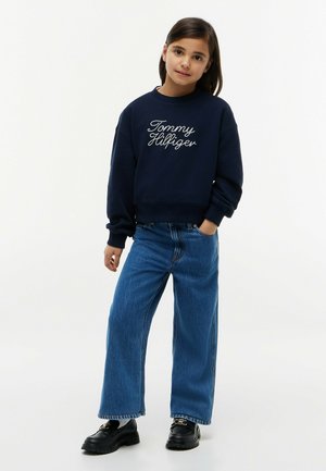Jeune fille aux cheveux foncés portant un sweat-shirt bleu marine Tommy Hilfiger, un jean bleu à jambes larges et des mocassins noirs, debout, une main dans la poche.