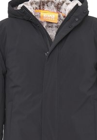 Giacca nera imbottita con cappuccio foderato in pelliccia sintetica, dotata di chiusura a zip e bottoni a pressione. Etichetta del marchio arancione sul rivestimento interno.