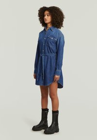 Τζιν φόρεμα, μπλε με ρυθμιζόμενη μέση, μακριά μανίκια, κλείσιμο με κουμπιά και μία τσέπη στο στήθος. Συνδυασμένο με μαύρες μποτες με πλατφόρμα.