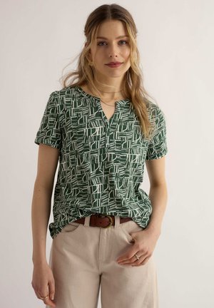 Joven mujer con cabello rubio ondulado, vestida con una blusa de manga corta con estampado verde, pantalones beige y un cinturón marrón, de pie frente a un fondo liso.