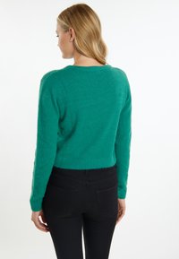 Maglione verde corto e sfocato con scollo rotondo e maniche lunghe, abbinato a jeans neri aderenti, che mostrano una superficie liscia.