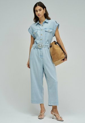 Mujer con mono sin mangas azul claro con cinturón, sosteniendo un bolso marrón claro y calzando tacones blancos de tiras frente a un fondo liso.