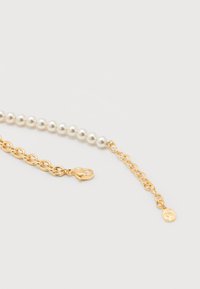 Χρυσό κολιέ αλυσίδα με λευκές πέρλες και κούμπωμα σε σχήμα αστακού σε λευκό φόντο, με μια μικρή στρογγυλή χρυσή ετικέτα με χαραγμένο το γράμμα "S".