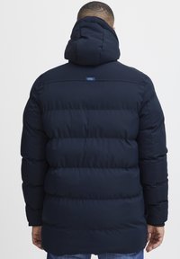Navy puffer coat with a hood, quilted design, and ribbed cuffs. Features a small label near the shoulder and a structured back panel.