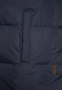 Surface d'une doudoune bleu marine avec une texture matelassée, présentant une ouverture de poche verticale et une étiquette de marque en cuir dans le coin inférieur droit.