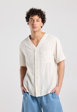 Jonge man met krullend haar, gekleed in een overhemd met korte mouwen, crème met strepen en lichtblauwe spijkershorts, met zijn handen in zijn zakken.