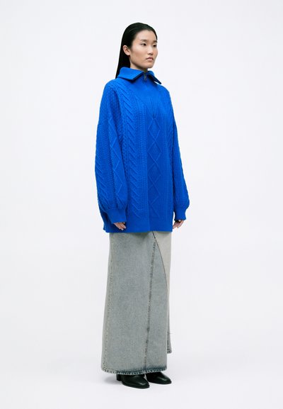 Suéter de punto azul con cuello alto, con patrones de cable texturizados, combinado con una falda maxi de denim gris y botas negras de tacón.