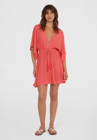 Koraalkleurige kaftan jurk met een V-hals, een cinched taille en losse, vloeiende mouwen. Gemaakt van een lichtgewicht stof met franjes.