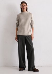 Beige gestrickter Pullover mit hohem Kragen, kombiniert mit dunkelgrauen, gestreiften, weit geschnittenen Hosen. Das Model steht vor einem hellen Hintergrund.