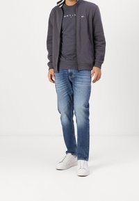 Grijze rits-hoodie met gestreepte binnenkraag, marineblauw T-shirt met logo, lichtblauwe jeans en witte sneakers. Casual outfit.