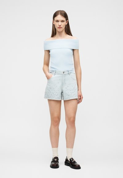 Γυναίκα με μακριά καστανά μαλλιά φοράει ανοιχτό μπλε τοπ με έξω τους ώμους, ανοιχτόχρωμα τζιν σορτς με στίγματα, μπεζ κάλτσες και μαύρα μοκασίνια, στέκεται μπροστά σε λευκό φόντο.