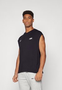Camiseta sin mangas negra de algodón, con un logo blanco en el pecho y un corte relajado. Combinada con pantalones de chándal grises.