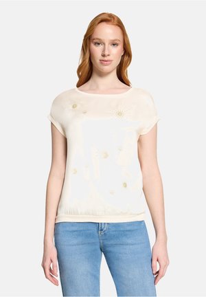 Frau mit roten Haaren, die eine elfenbeinfarbene Kurzarmbluse mit dezenten goldenen Blumendesigns und hellblaue Jeans trägt, steht vor einem weißen Hintergrund.