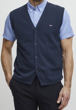 Man wearing a navy blue buttoned vest over a light blue short-sleeve collared shirt and black pants, with hands in pockets.