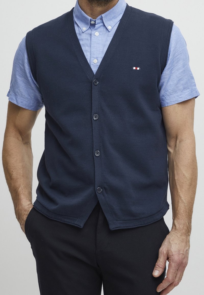 Homme portant un gilet bleu marine boutonné par-dessus une chemise à manches courtes bleu clair avec col, et un pantalon noir, les mains dans les poches.