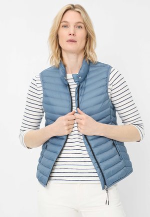 Blonde Frau, die eine hellblaue gesteppte Weste über einem weiß-navy gestreiften langärmligen Shirt vor einem schlichten hellen Hintergrund schließt.