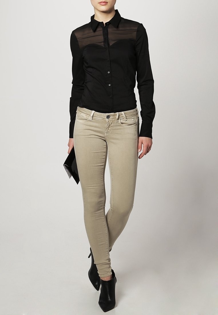 Femme portant un chemisier noir boutonné avec un détail transparent en haut, un pantalon beige slim et des bottines noires, tenant une pochette noire dans sa main gauche.