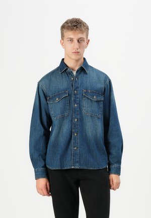 Young man standing, wearing a blue denim button-up shirt with two chest pockets and black pants against a plain white background.