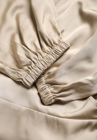 Tessuto beige con una texture liscia, caratterizzato da polsini elastici arricciati e una leggera lucentezza. Il design include morbide pieghe e un aspetto drappeggiato.