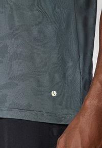 Σκούρο πράσινο αθλητικό μπλουζάκι με υφή, παραλλαγής και μικρό λευκό λογότυπο κοντά στο στρίφωμα. Κοντινή λήψη του υφάσματος και λεπτομερειών.