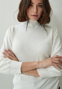 Balta ribota megztinė su pūstomis ilgomis rankovėmis, turinti prigludusią ragą ir tekstūruotą audinį, dėvima su sukryžiuotomis rankomis.