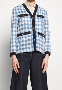 Blazer in tweed con motivo a quadri blu e bianchi, con rifiniture nere, bottoni e due tasche frontali. Il tessuto ha una finitura testurizzata.